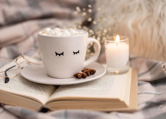 Tasse de chocolat chaud avec des marshmallows, posée sur un livre ouvert, accompagnée d’une bougie et d’une ambiance cosy.