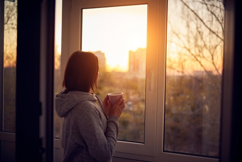 Femme de dos regardant le lever du soleil par la fenêtre, une tasse à la main, moment de calme et de réflexion.