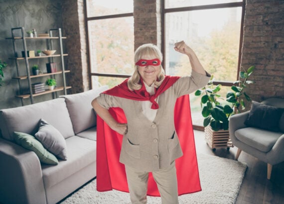Femme âgée souriante déguisée en super-héroïne, debout dans un salon, symbole de force discrète et d’affirmation de soi.