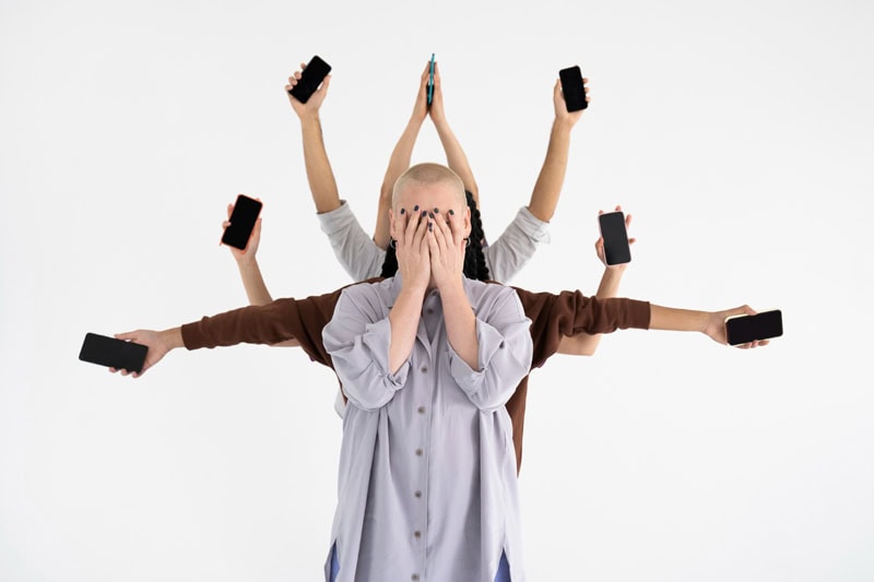 Personne les mains sur le visage, entourée de bras tenant des smartphones, symbole de la pression numérique et du trop-plein sur les réseaux sociaux.