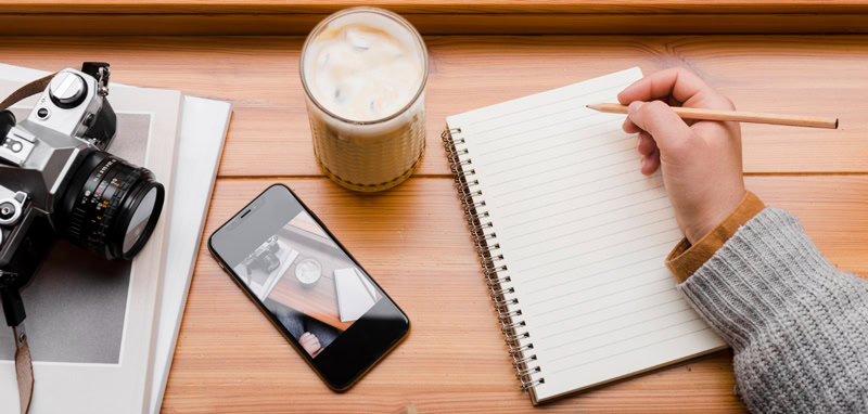 Main écrivant dans un carnet à côté d’un téléphone, d’un appareil photo et d’un café glacé, symbole d’un choix conscient entre création personnelle et exposition numérique.