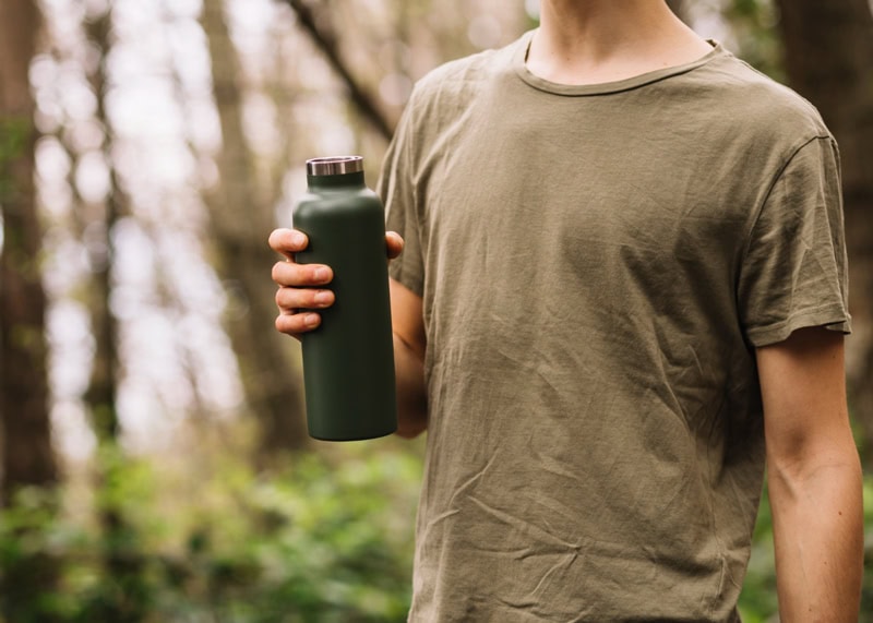Personne tenant une gourde verte en métal lors d’une balade en forêt, symbole d’aventure et d’hydratation.