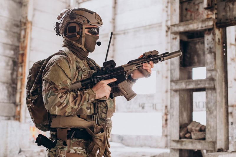 Personne en tenue militaire camouflée tenant une arme dans un bâtiment abandonné.