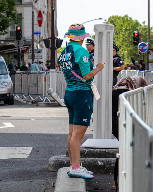 Bénévole des Jeux de Paris 2024 en tenue officielle turquoise, debout dans une rue parisienne près de barrières de sécurité, avec un panneau et une bouche de métro en arrière-plan.