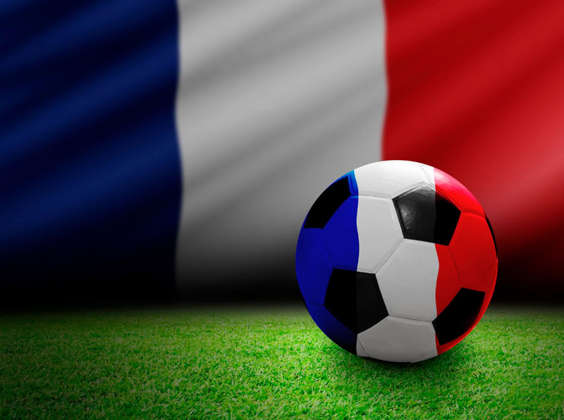 Ballon de football aux couleurs du drapeau français posé sur un terrain de gazon, avec un grand drapeau bleu blanc rouge en arrière‑plan.