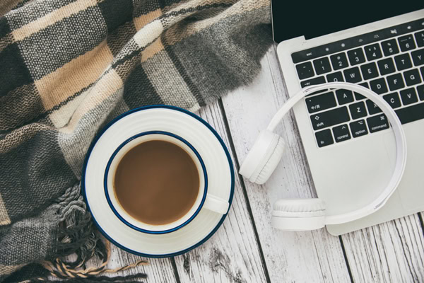 Ordinateur portable avec un casque audio, une tasse de café et un plaid à carreaux sur une table en bois blanc.