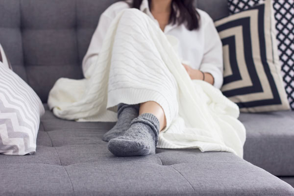 Personne installée sur un canapé gris, les pieds dans de grosses chaussettes et enveloppée dans un plaid blanc.