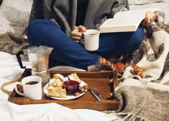 Personne lisant un livre sous une couverture, avec des tasses de café et un plateau gourmand.