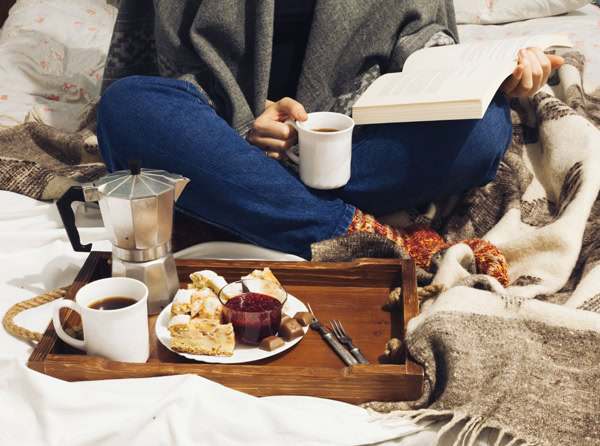 Personne lisant un livre sous une couverture, avec des tasses de café et un plateau gourmand.